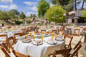 Outdoor wedding reception setup with white tablecloths and wooden chairs under string lights.