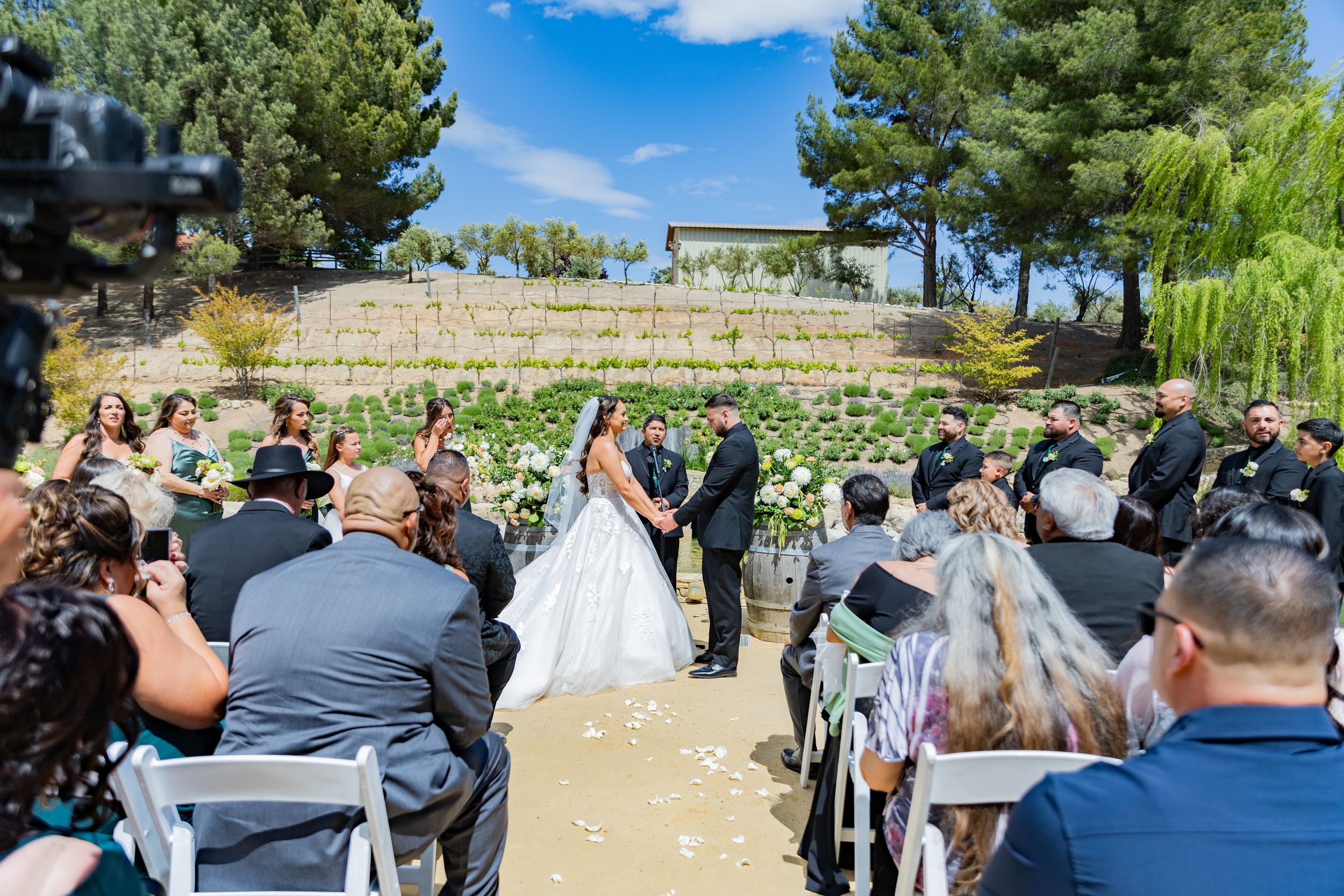 Wedding ceremony taking place outdoors with guests seated and stone terraces in the background.