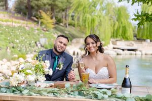 Wedding couple enjoying drinks at decorated table by scenic pond.