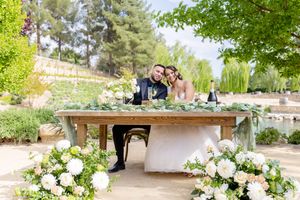 Couple seated at elegantly decorated table with white floral arrangements in garden setting.