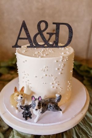 White wedding cake with black monogram topper and small dog figurines as decorations.