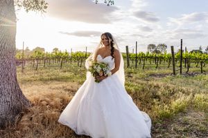 Bride in flowing white gown poses in vineyard at sunset.