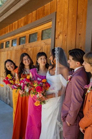 Wedding party in colorful orange and pink dresses gather by wooden cabin wall.