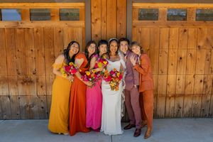 Wedding party with bridesmaids in bold orange and pink dresses pose against wooden building.