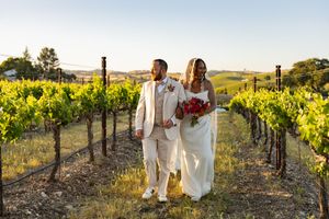 Couple in light colored wedding attire walking through scenic vineyard rows at sunset.
