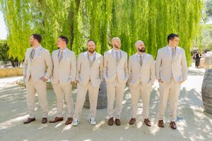 Six groomsmen wearing matching light beige suits pose under a weeping willow tree.