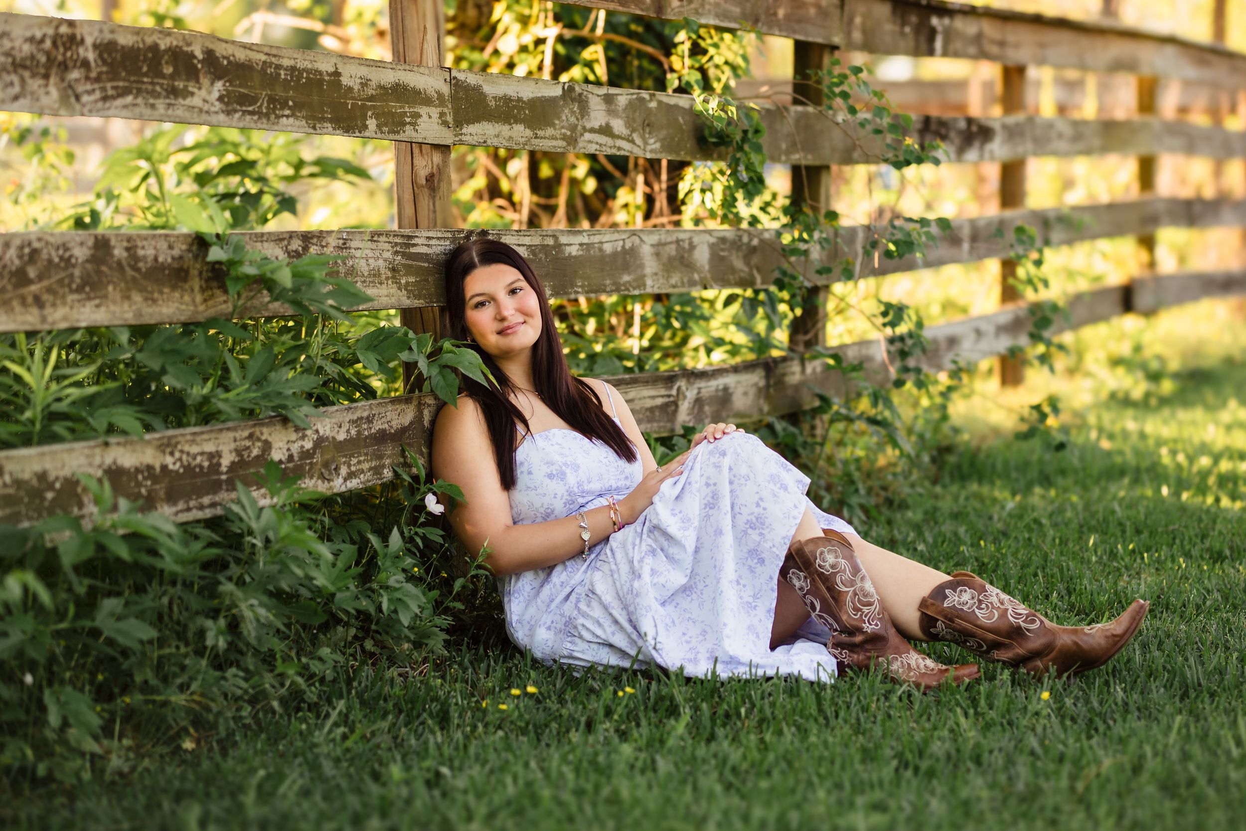 Person in white dress poses on grass near rustic wooden fence at sunset.