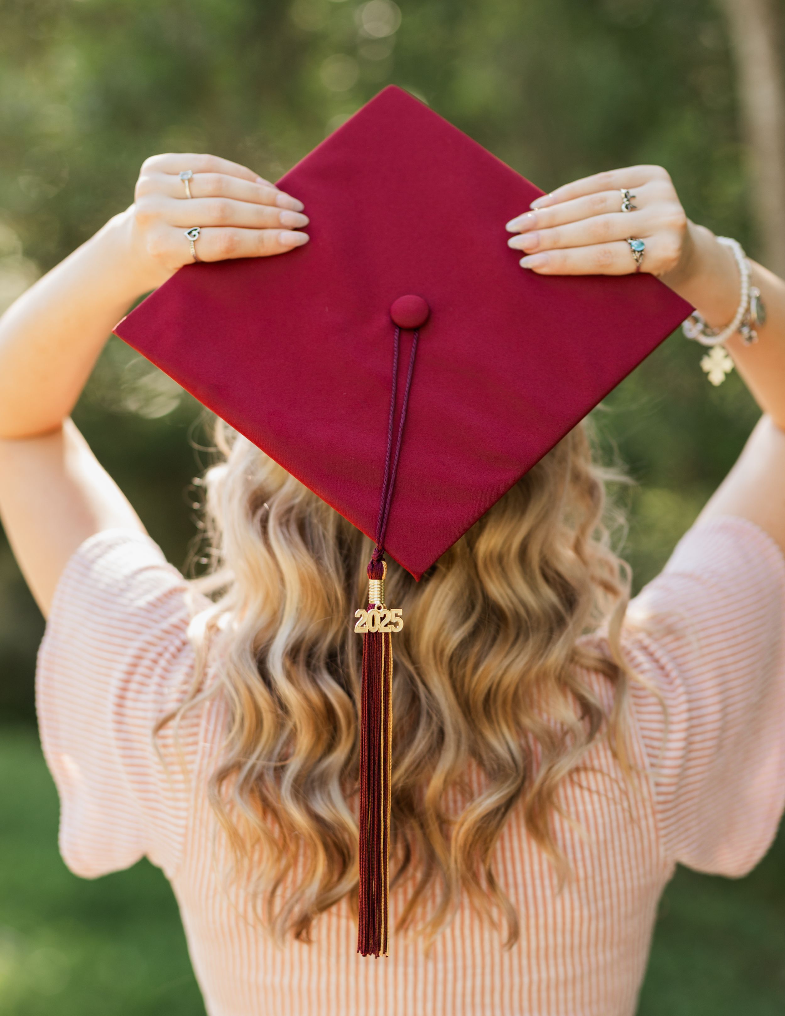 Graduate holds up burgundy cap while wearing pink sweater in outdoor graduation photoshoot.