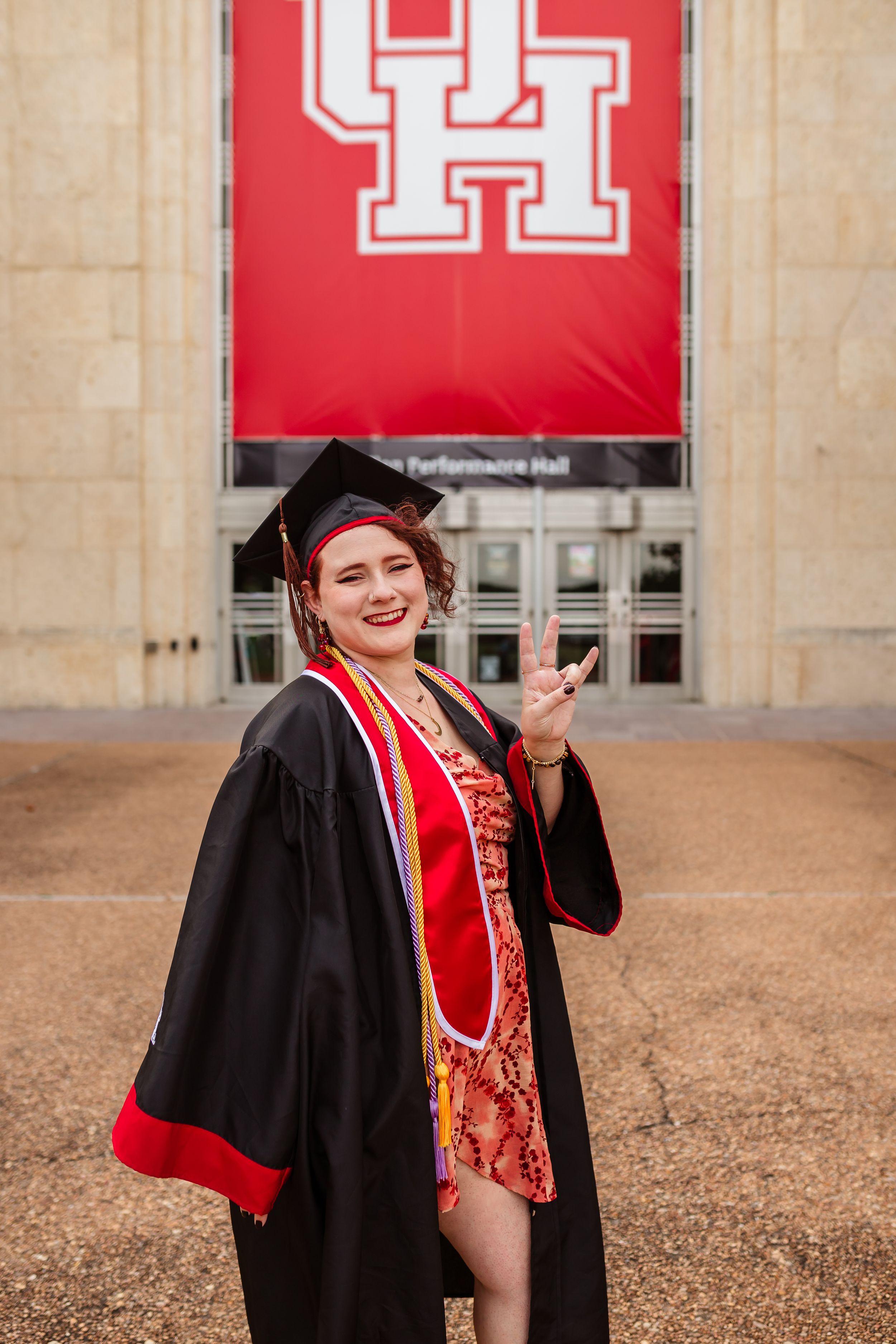 Graduate in black cap and gown with red sash poses joyfully in front of University of Houston building with red banners.