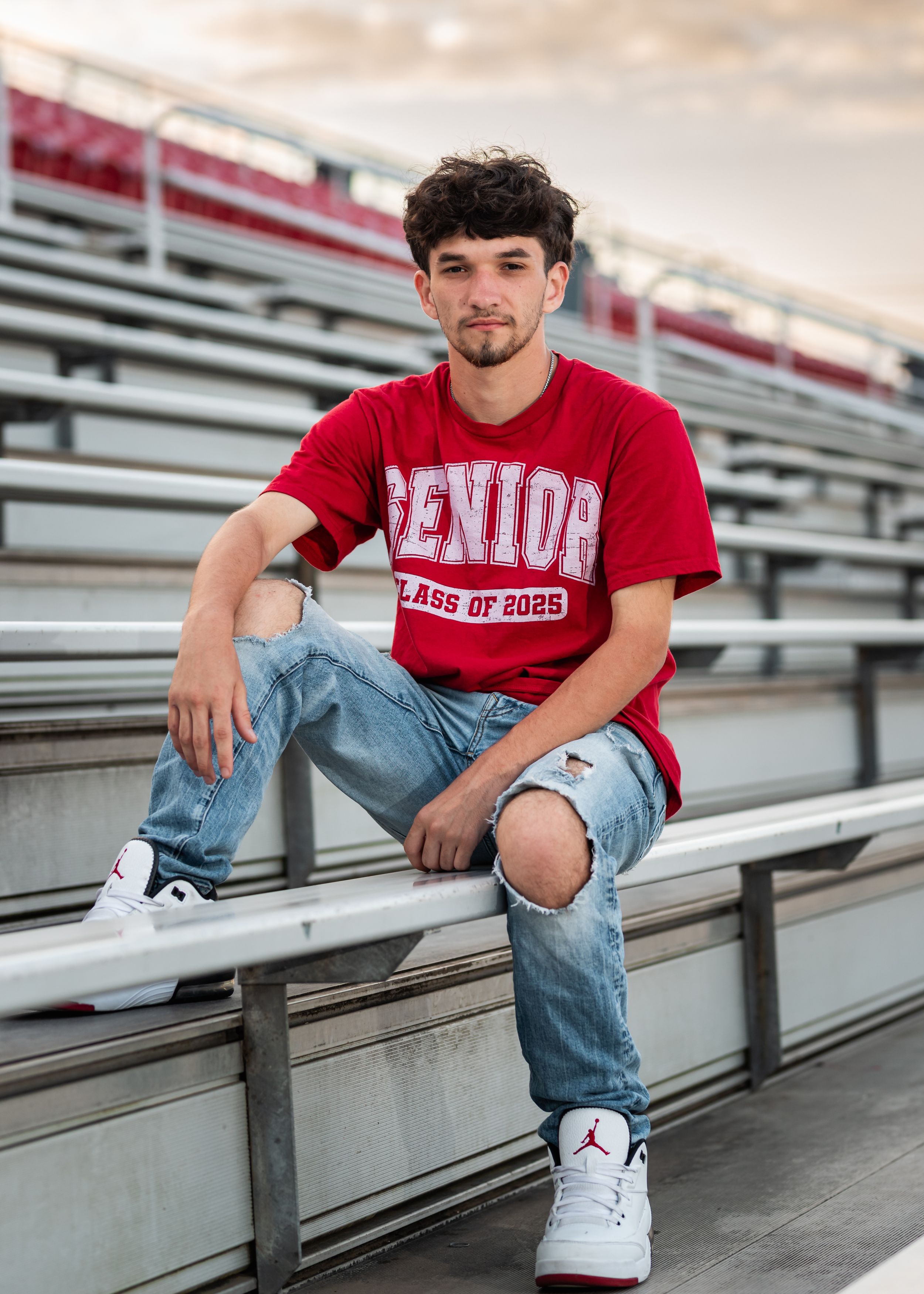Person in red shirt and jeans poses on stadium bleachers wearing white sneakers.