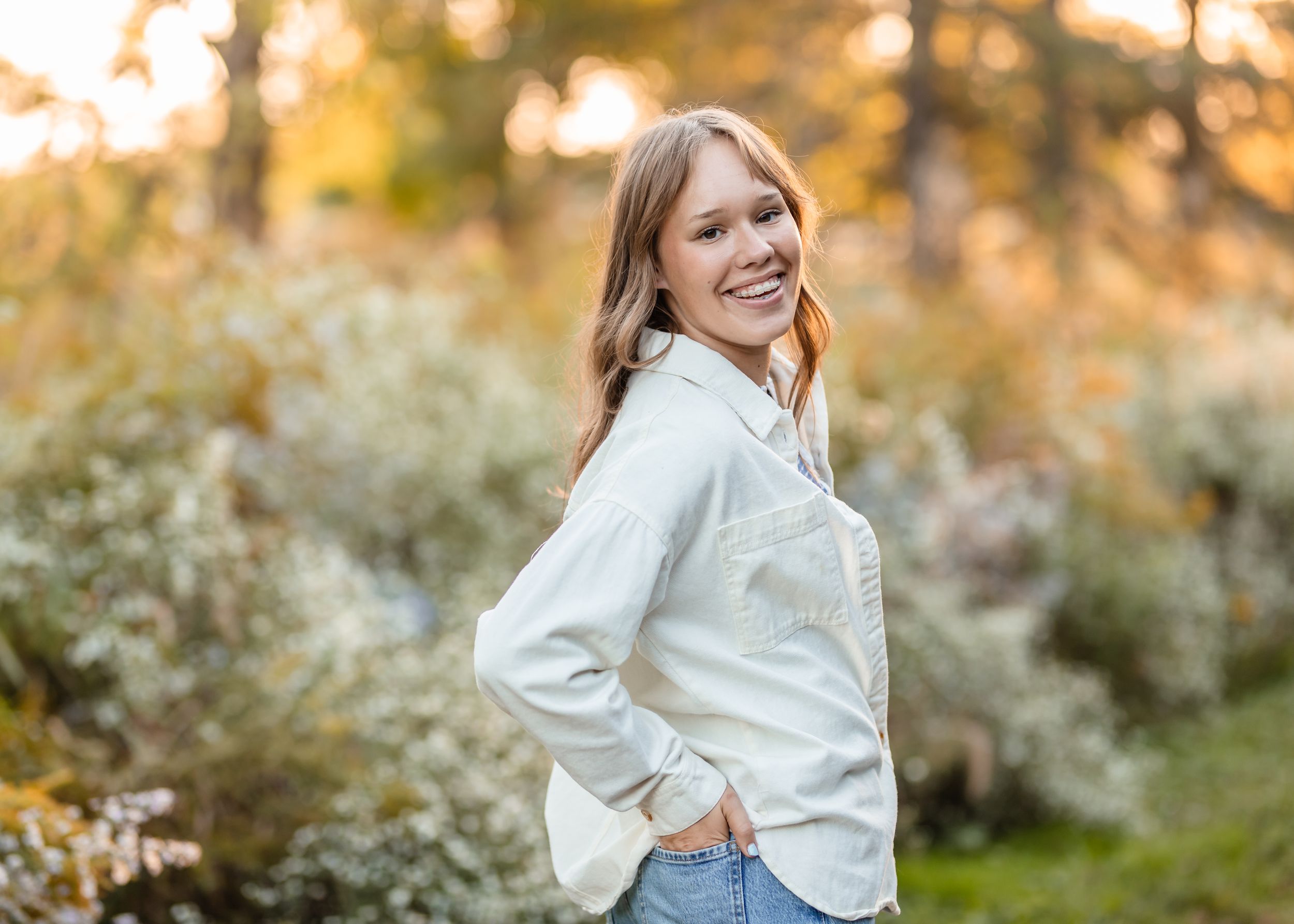 A high school senior in white button-down shirt and jeans poses outdoors during golden hour with blurred foliage background.