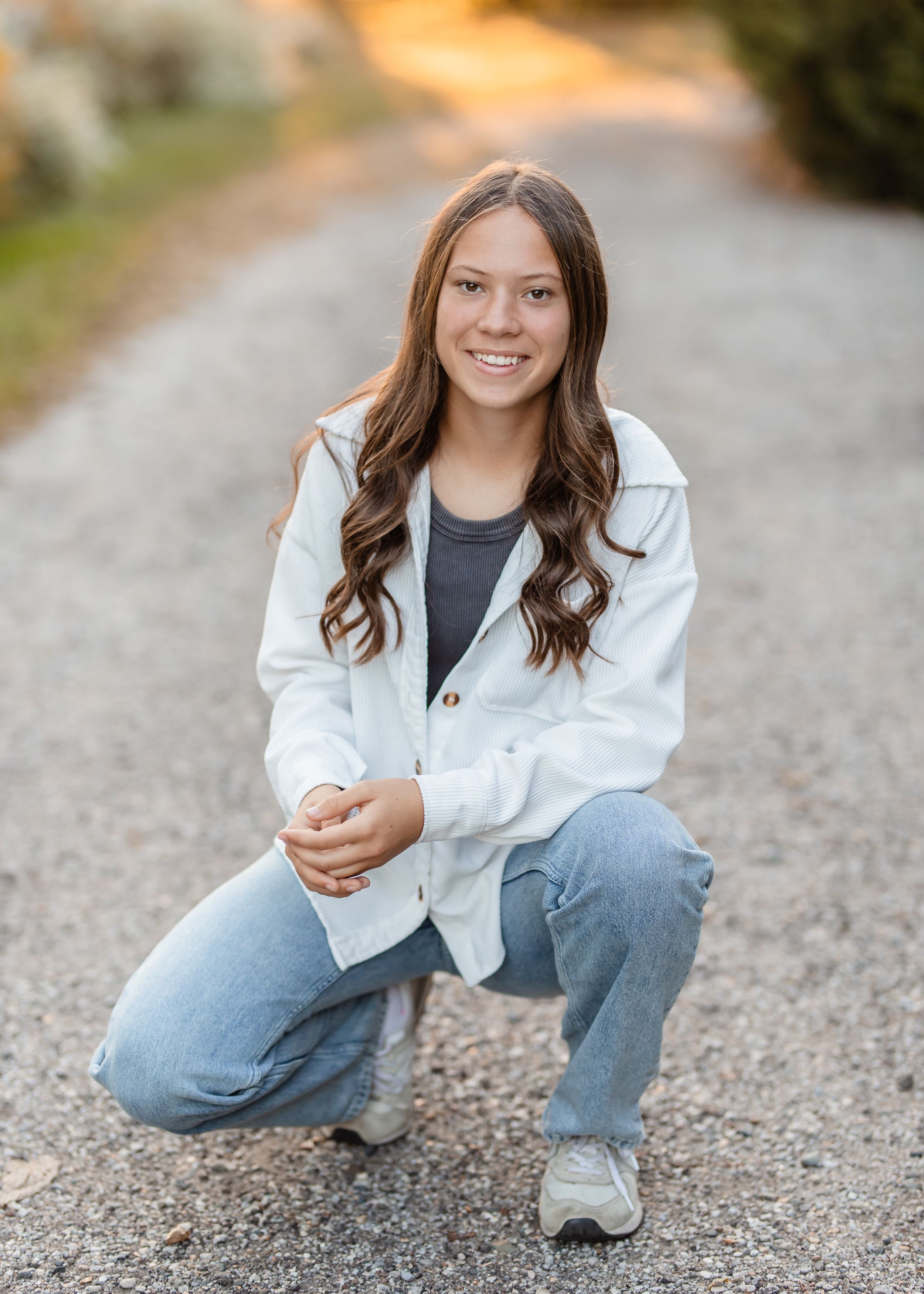 A person in a white jacket and blue jeans crouches on a gravel path during an outdoor senior photo session.