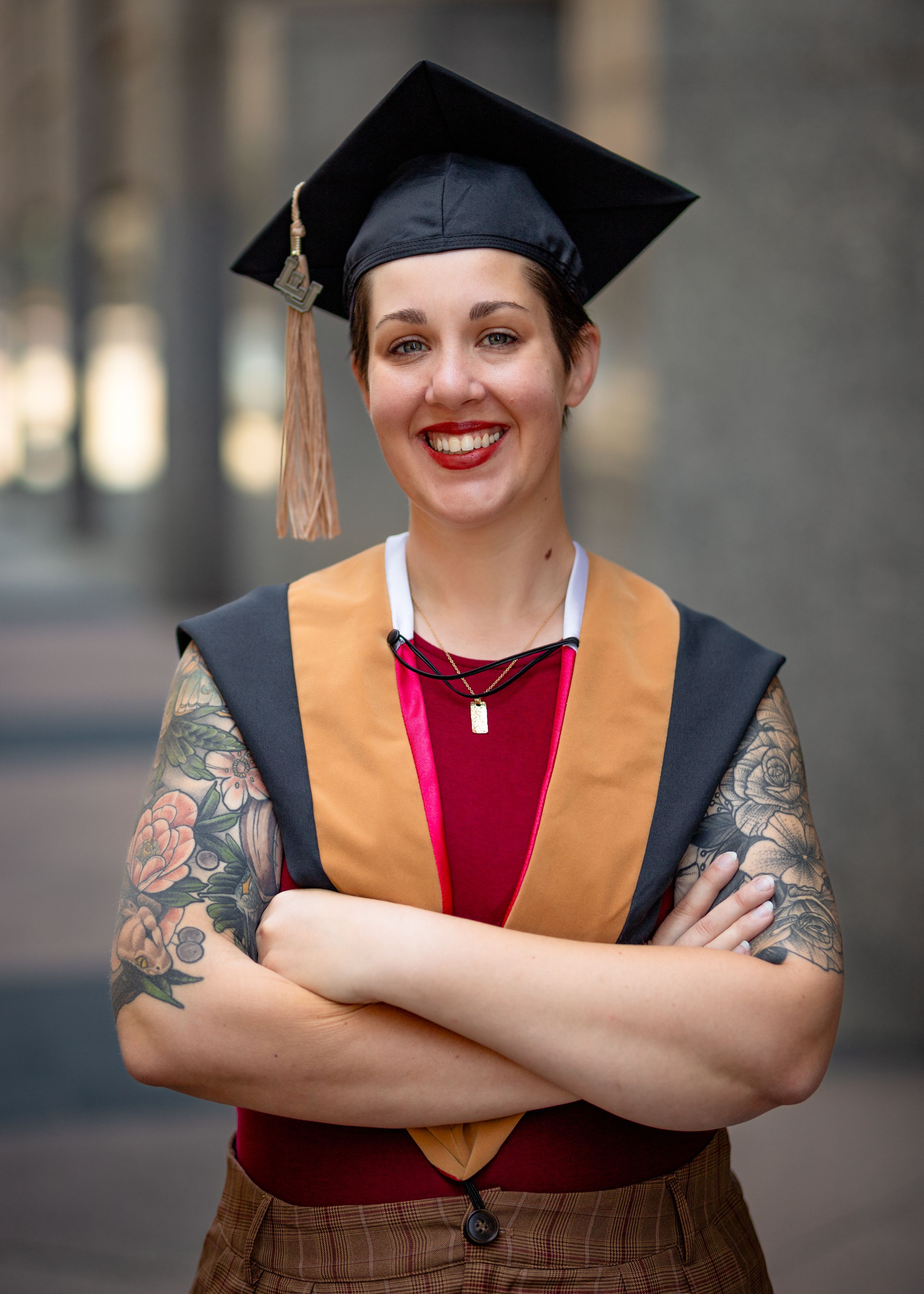Graduate in cap and gown with gold stole shows tattoos while posing confidently.