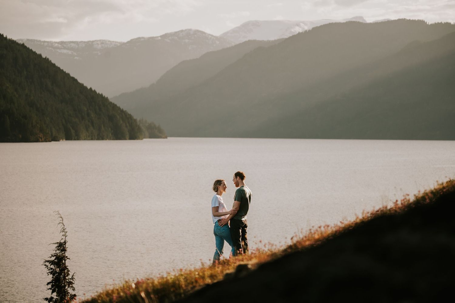 Golden hour engagement session at Comox Lake, couple embracing near rocky cliffs on Vancouver Island in front of Comox Valley Glacier