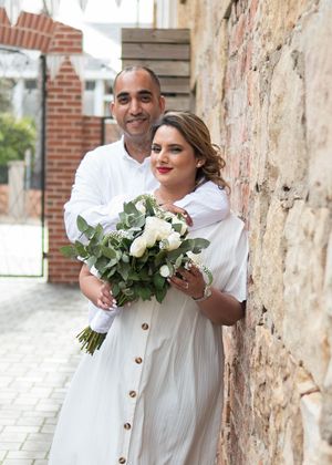 A happy couple in wedding attire poses together against a rustic stone wall while holding a beautiful white floral bouquet.