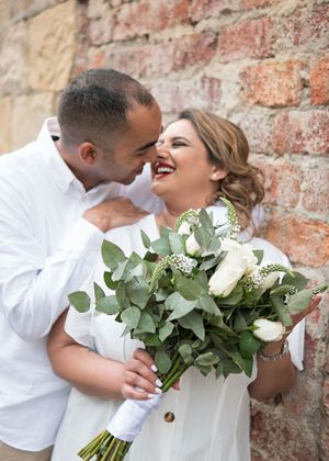 A joyful wedding moment captured against a rustic brick wall with a bride holding a white and green eucalyptus bouquet.