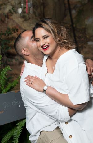 A romantic couple wearing white outfits share a joyful moment together outdoors near green ferns.