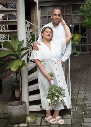 A couple dressed in white outfits stand together on outdoor stairs near a potted plant and ornate railing.