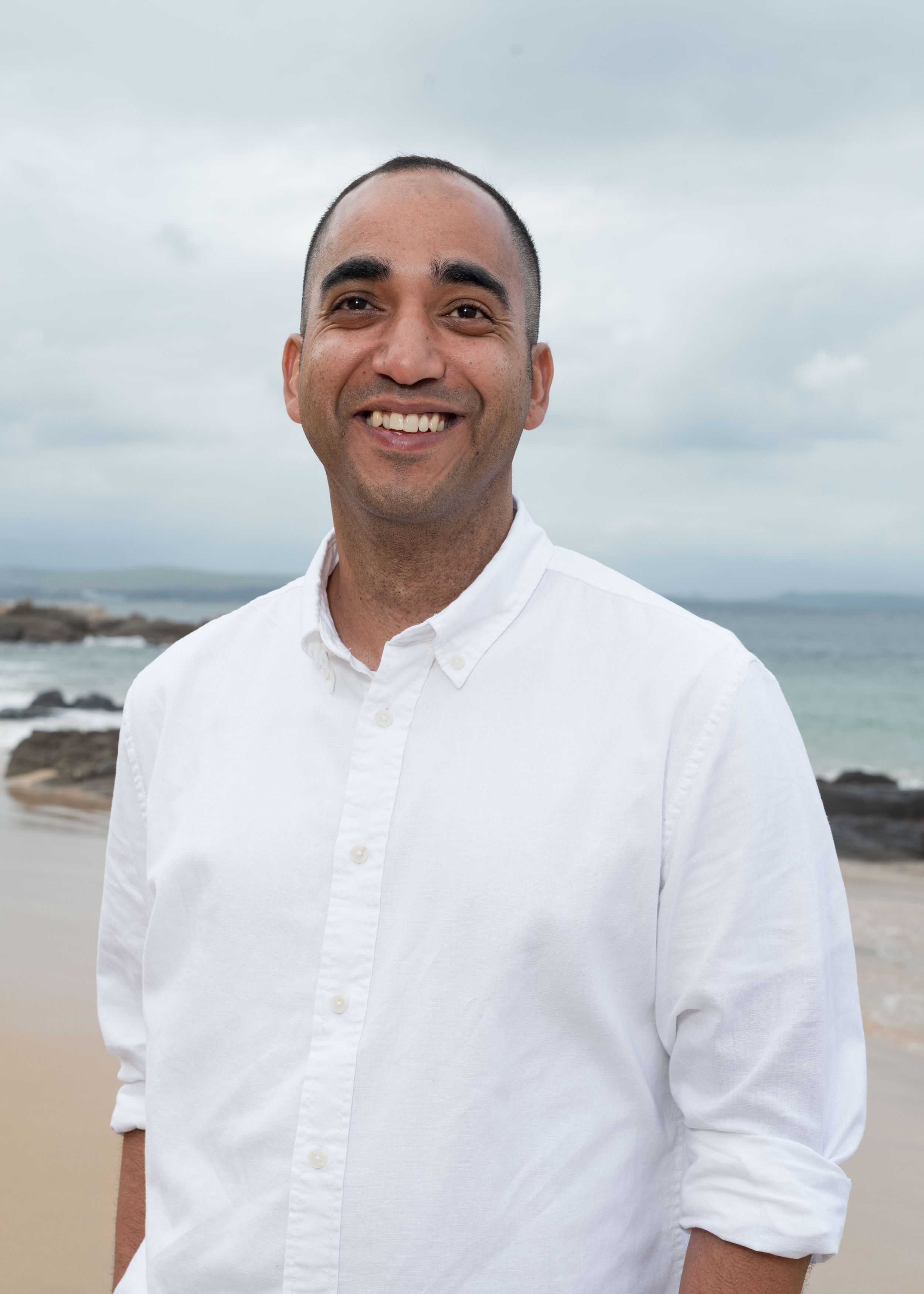 Person wearing a white button-down shirt smiles at the camera while standing at a rocky beach on an overcast day.