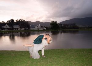 A couple embraces in a dip pose on grass beside a tranquil lake at sunset with mountains in the background.