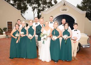 Bridal party poses in front of white church with bridesmaids wearing emerald green dresses and holding bouquets.