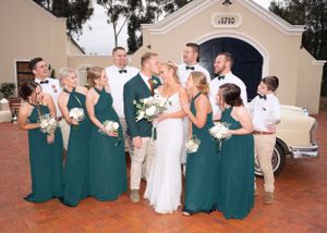 Wedding party in emerald green bridesmaid dresses and khaki suits gather on brick patio during outdoor celebration.