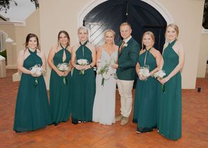 Wedding party poses in matching emerald green dresses and khaki suit against arched doorway at venue.