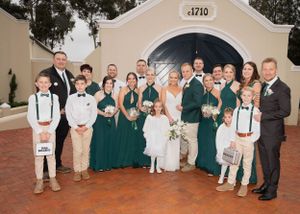 Wedding party wearing emerald green and neutral colors poses together on brick patio in front of building entrance.