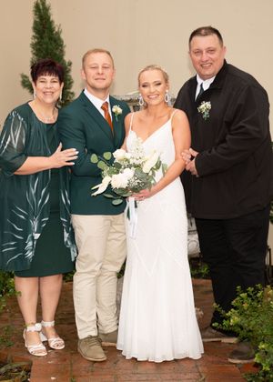 Small wedding group stands together near bushes and entrance with bride in white dress and others in green and black attire.