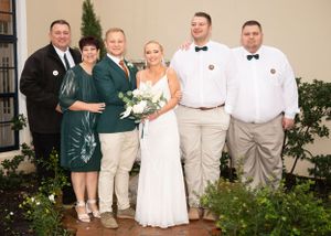 A group celebrates at a wedding ceremony wearing white, tan and green attire while posing in a garden setting.