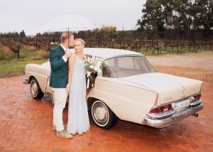 Couple poses next to a vintage cream-colored Mercedes sedan on brick pavement at a countryside wedding venue.