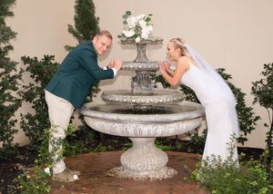 Couple in formal attire playfully posing at an ornate stone fountain decorated with white flowers and greenery.