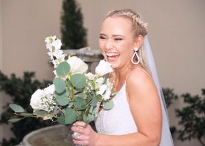 A joyful bride in a white wedding dress laughs while holding a bouquet of white flowers and eucalyptus.