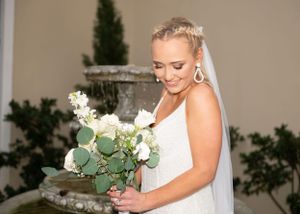 A bride in a white wedding dress holds a beautiful bouquet of white flowers and eucalyptus leaves near a fountain.