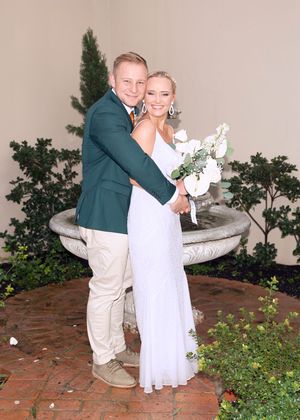 A couple poses together at a garden wedding venue with brick pavers and an ornate stone fountain in the background.