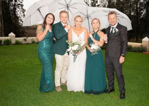 A wedding party poses together under a clear umbrella on a rainy day on a green lawn.