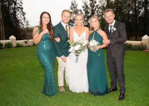 Wedding party in emerald green dresses and suits pose together on manicured lawn at outdoor venue.