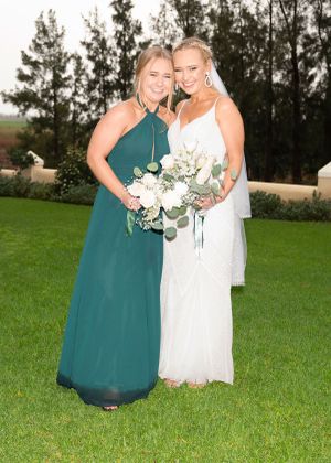 Two people pose together in formal dresses, one wearing emerald green and one in white lace, holding floral bouquets on a lawn.