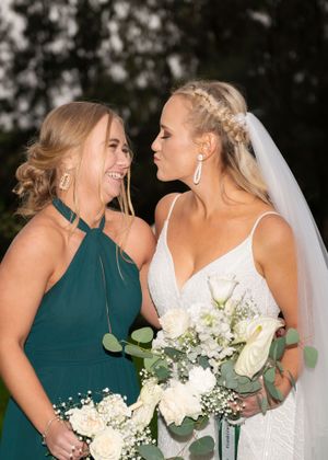 Two women in formal attire share a joyful moment at a wedding, one wearing emerald green and one in a white bridal gown.