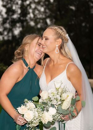 Bride with braided hair and veil shares joyful laughter with bridesmaid in emerald dress while holding white bouquet.