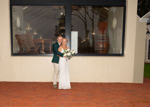 Two people embrace in a sweet moment outside a building with large windows and rocking chairs visible inside.