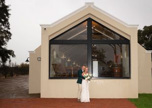 Couple embraces outside a modern white chapel building with large glass windows on a rainy day.