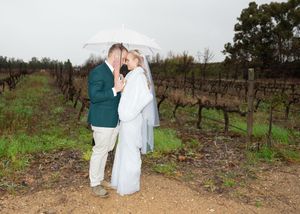A couple embraces under a white umbrella at a vineyard on a cloudy day.
