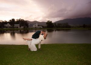 Romantic wedding dip pose on grass by a serene lake at sunset with mountains in the background.