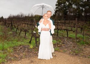 A bride in white dress holds a white umbrella while standing on a dirt path in front of vineyard rows.