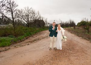 A couple celebrates their wedding day walking together on a rustic dirt road surrounded by green trees on a cloudy day.