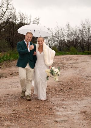 A couple in wedding attire walks down a dirt path under a white umbrella on an overcast day.