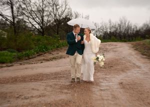 A couple shares an umbrella while walking down a muddy rural road in wedding attire on an overcast day.
