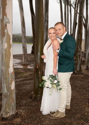 A romantic wedding photo showing a couple embracing among eucalyptus trees, with the bride holding a white floral bouquet.