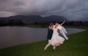 A couple dances joyfully by a lake beneath stormy skies and mountains in a romantic outdoor scene.